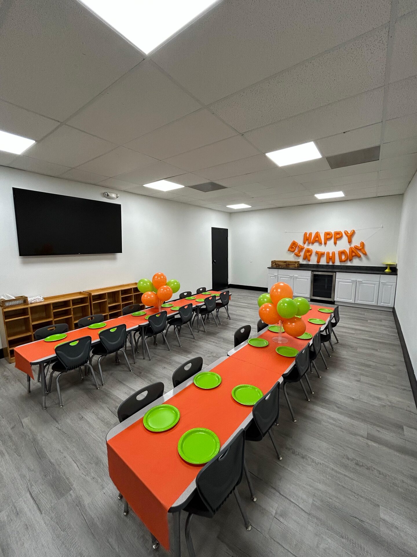 Birthday party setup in the Cedar Park event room with long orange tables, green plates, balloons, and a Happy Birthday banner
