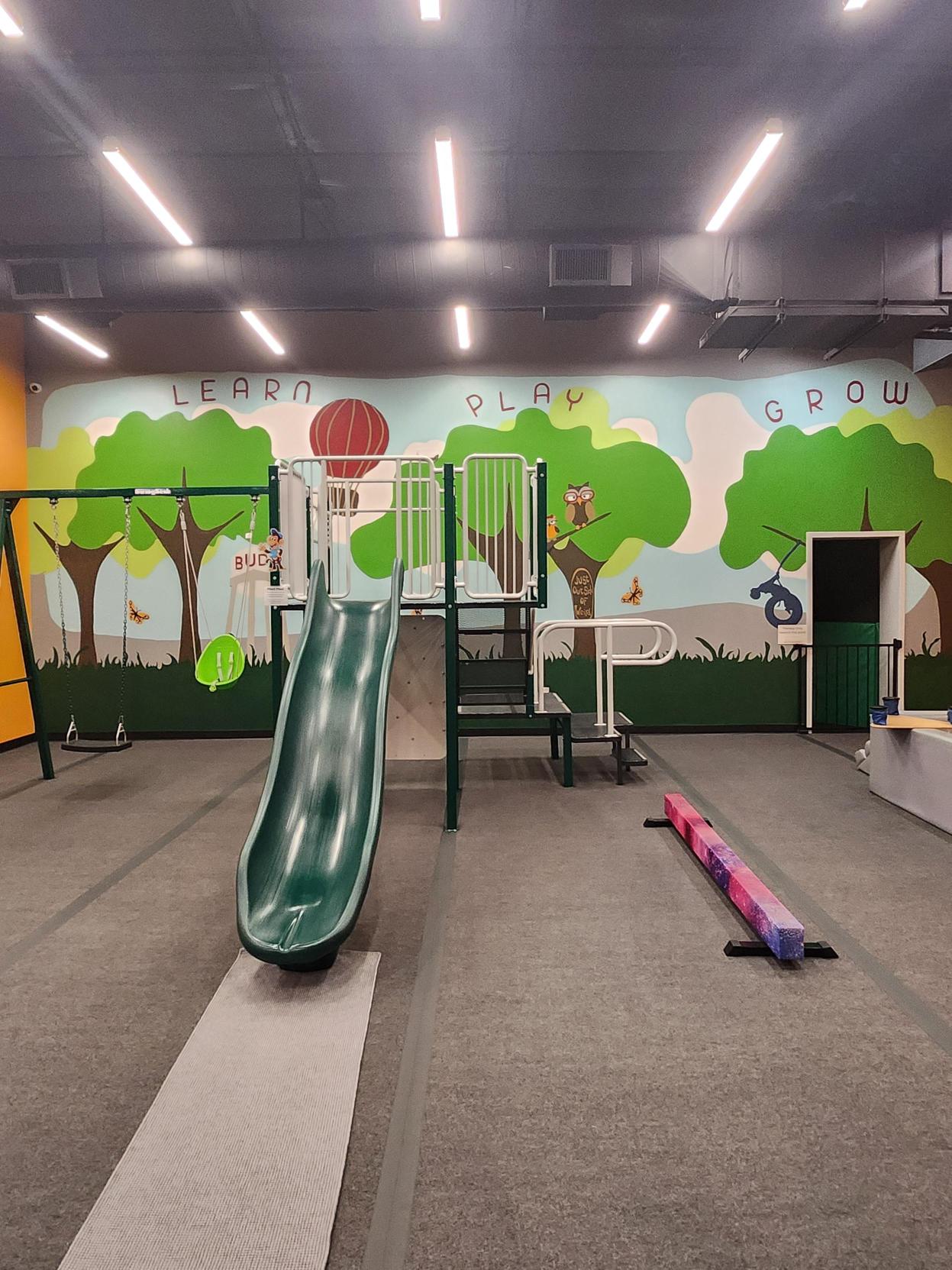 Indoor playground with green slide and hand-painted 'Learn Play Grow' tree mural at Little Land Buda