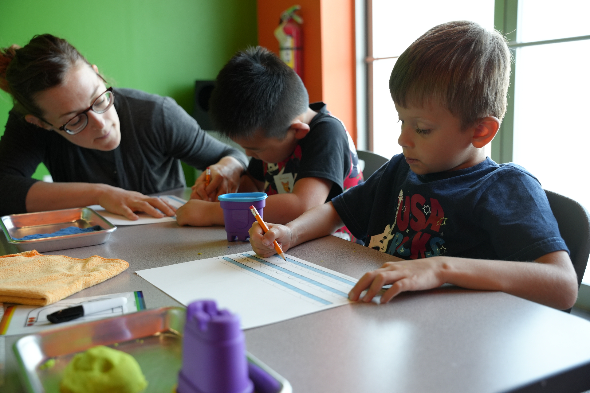 Teacher helping two boys with handwriting practice at a sunny classroom table